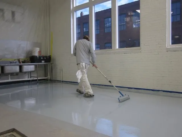 Installer rolling an epoxy coating across a floor beside tall windows